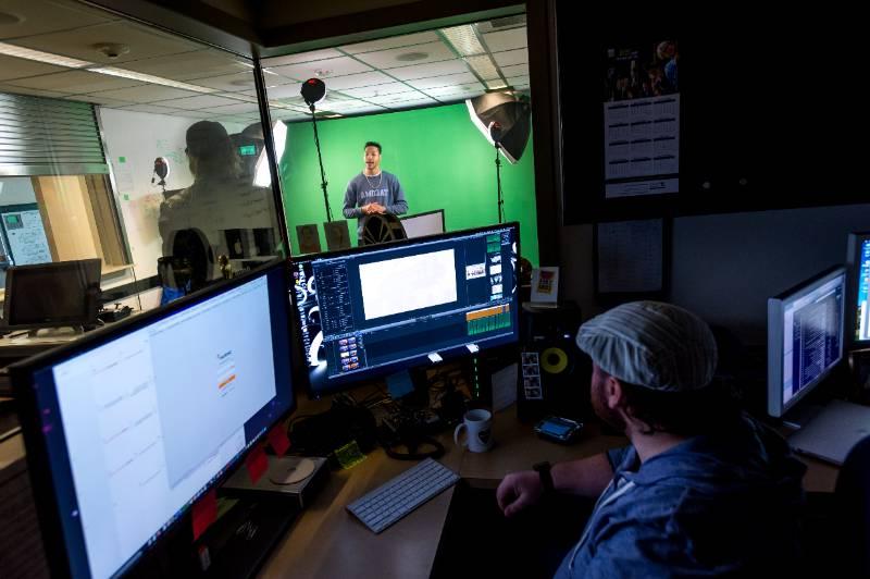 A student stands in front a green screen and video set up, while another edits footage on multiple screens